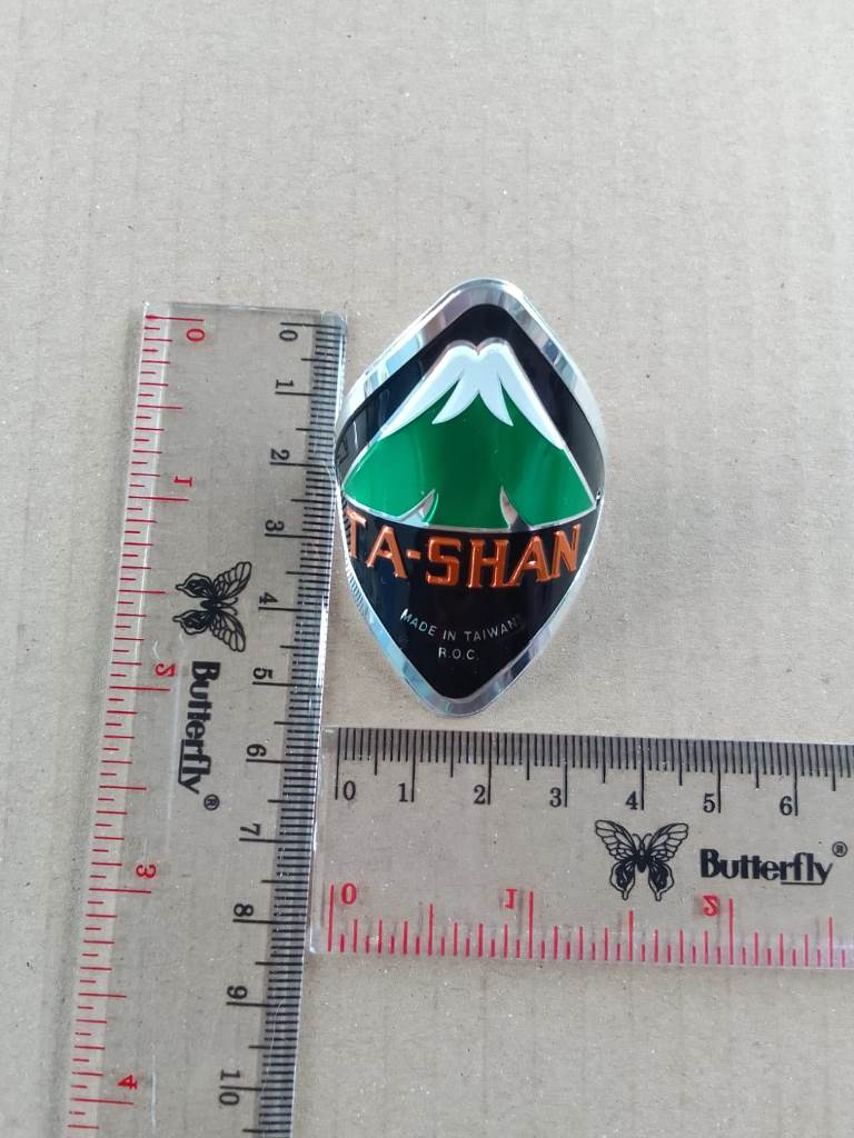 Metallic badge with 'Ta-Shan' logo next to a ruler on a gray background